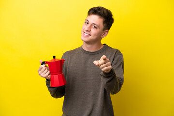 Young Brazilian man holding coffee pot isolated on yellow background pointing front with happy expression