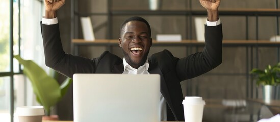 Young businessman rejoicing with raised arms at his desk, gazing at laptop screen.
