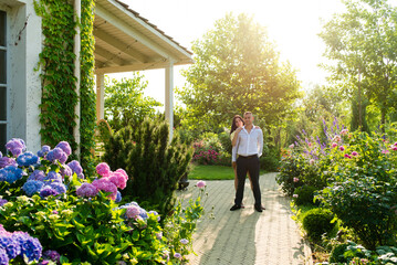 Happy couple in love walks and cuddles in the garden near the house. Well-groomed lush green summer garden with flowers.