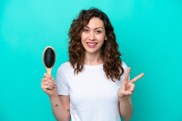 Young caucasian woman holding hairbrush isolated on blue background smiling and showing victory sign - obrazy, fototapety, plakaty