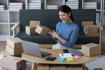 Young Asian woman working with parcel box at home preparing to deliver goods and writing online order note from internet on laptop.