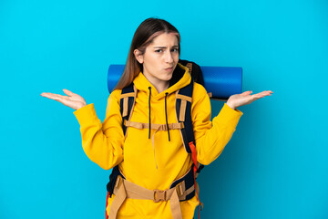 Young mountaineer woman with a big backpack isolated on blue background having doubts while raising...