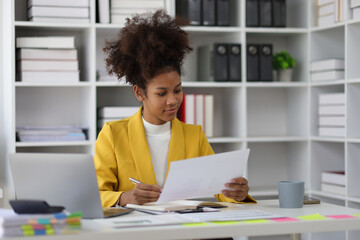 Attractive african american businesswoman working with laptop and documents on office desk writing work notes on notepad.
