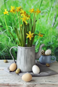 Easter eggs and a bouquet of daffodils in a jug, on a wooden table, against a background of a green lawn, Easter still life