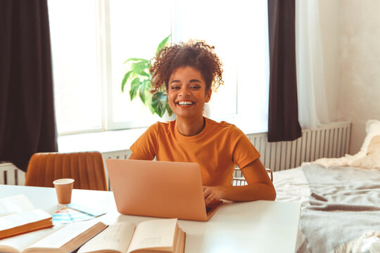 Cheerful young Afro American female student studying online, sitting at desk surrounded by textbooks