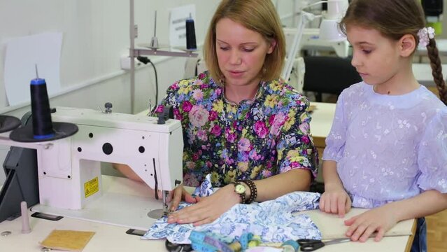 tailor sits at table with sewing machine and shows seam to girl