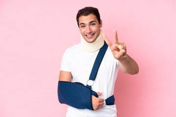 Young caucasian man with broken arm and wearing a sling isolated on pink background smiling and...