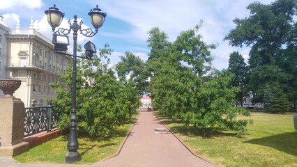 In the park, trees grow on the lawns and metal poles with lanterns are installed near the tile...