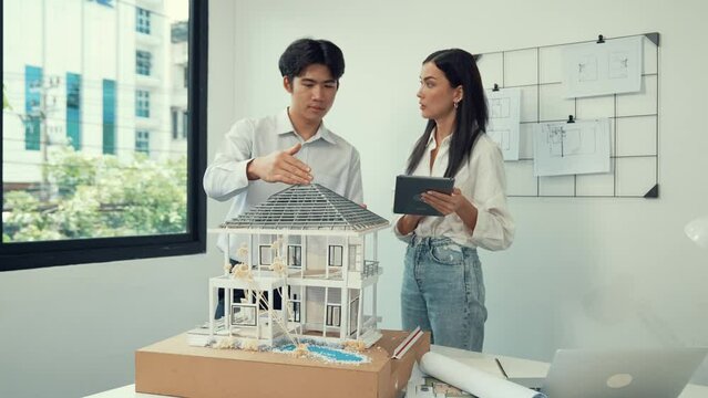 Beautiful young caucasian project manager brief asian engineer by using tablet while engineer measuring house model on meeting table with blueprint, architectural equipment scatter around. Immaculate