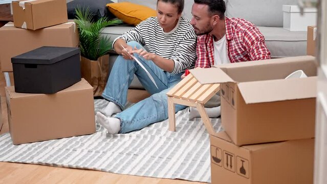 Home DIY Delight: A joyful young couple assembles modern wooden shelf furniture amidst moving boxes, infusing their new apartment with DIY home decoration
