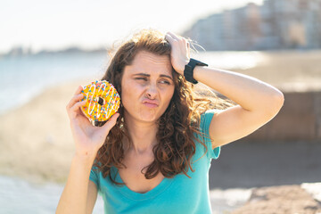 Young sport woman holding a donut at outdoors having doubts and with confuse face expression