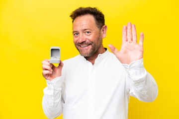 Middle age man holding a engagement ring isolated on yellow background saluting with hand with...