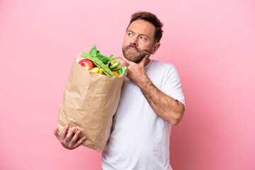 Middle age man holding a grocery shopping bag isolated on pink background having doubts
