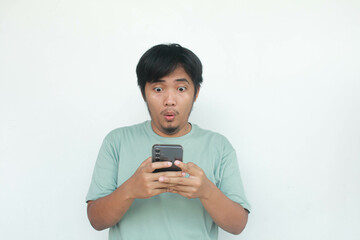 Portrait of a young Asian man in a green t-shirt casually holding his cellphone with a happy and wow expression. Isolated on gray background.