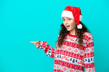 Little caucasian with christmas hat isolated on blue background surprised and pointing finger to...