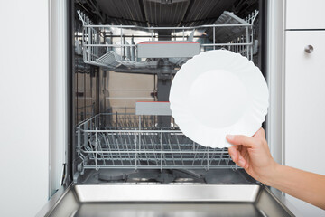 Young adult woman hand holding and showing white clean plate on dishwasher background at home...