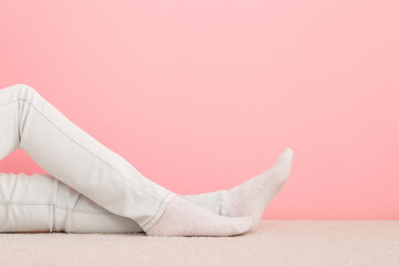 Girl long legs in white jeans and socks sitting on light beige carpet at pink wall background. Pastel color. Closeup. Side view. Empty place for text.