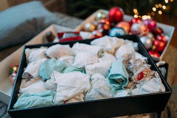 In the foreground there is a lot of colorful paper for packing Christmas tree glass toys to hide them for storage. In the background there is a Christmas toys with blurred yellow lights and garlands