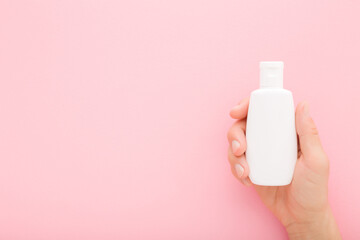Young adult woman hand holding white plastic cream bottle on pink table background. Pastel color. Care about clean and soft body skin. Female beauty product. Closeup. Empty place for text. Top view.