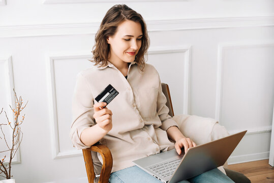 Happy millennial woman typing on laptop, seamlessly using credit card for online shopping, creating a modern image of convenience and enjoyment of digital retail experience at home.
