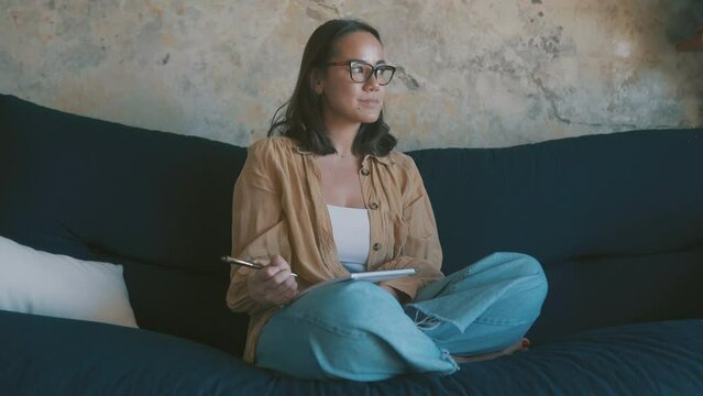 Thinking, writing in notepad and woman on sofa at home for creative idea, planning and schedule. Inspiration, notes and Asian person on couch with to do list, journal and pen to study in living room