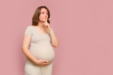 A pregnant woman with her finger on her lips on a pink studio background. Pregnancy in a woman with...