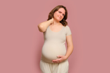 A pregnant woman holds onto a sore neck on a studio pink background. Pregnancy in a woman with a belly
