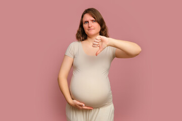 A pregnant woman shows a disapproving thumbs-down gesture on a studio pink background. Pregnancy in...