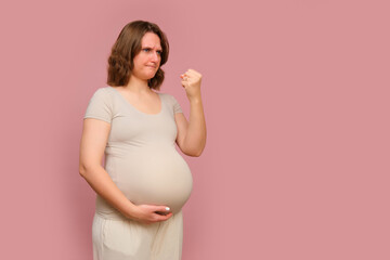 A pregnant woman showing her fist on a pink studio background. Pregnancy of an angry woman with a belly, copy space