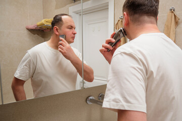 A man shaves with an electric razor in the bathroom in front of the mirror. Overweight 40-year-old...
