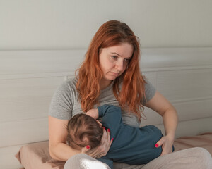 An unhappy woman breastfeeds her son and sits on the bed. Postpartum depression.