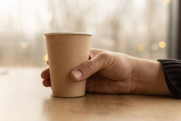 A hand holding a paper cup on the table near the window, blurred background. Mockup, copy space