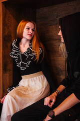 A girl with flowing red hair in a black blouse with ruffles and a long white skirt sits in the...