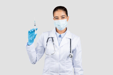 Confident female medical professional in a white coat and face mask holds a syringe