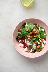 Feta cheese, baby spinach, pomegranate and almonds salad in a roseate bowl, above view on a light-beige stone background, vertical shot
