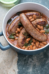 Serving pan with roasted sausages and borlotti beans, vertical shot on a grey and beige granite background, middle close-up, selective focus