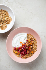 Granola bowls with yogurt, pomegranate seeds and nuts, above view on a light-beige stone background, vertical shot with space