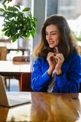 romantic young woman in blue shirt making a heart gesture with her fingers in front of her chest In front of her laptop showing her love and affection, concept St. Valentine's Day