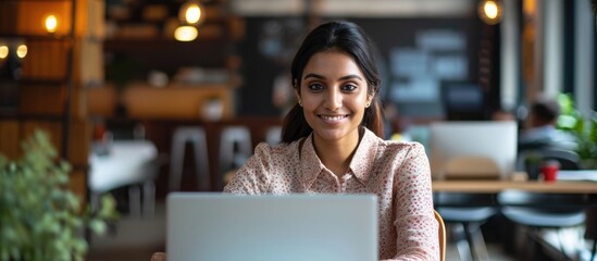 Happy and accomplished female Indian programmer working in a contemporary office, providing tech support by typing on her laptop with a smile. - obrazy, fototapety, plakaty