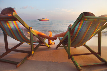 Happy couple sitting at sun chairs on the beach of Koh Samet at sunset