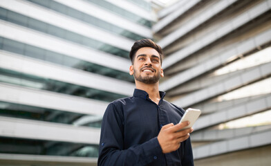 Confident middle eastern young businessman holding cellphone near office building