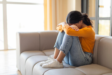 Emotionally overwhelmed indian woman sitting on couch, covering her face with arms