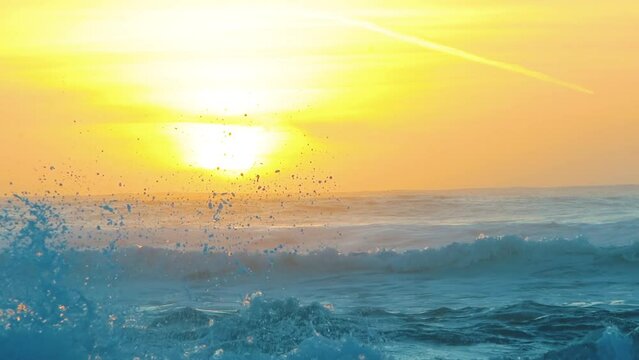 A slow motion shot of the sun setting on the horizon while som beautiful crystal blue waves are crashing on the foreground.