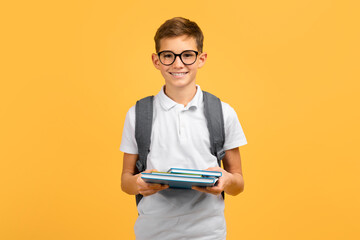 Happy teen boy wearing glasses and backpack holding workbooks - obrazy, fototapety, plakaty