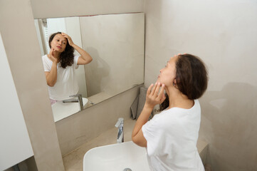 Happy young woman touching her face, examining flaws, skin problems and wrinkles, standing in front of a bathroom mirror