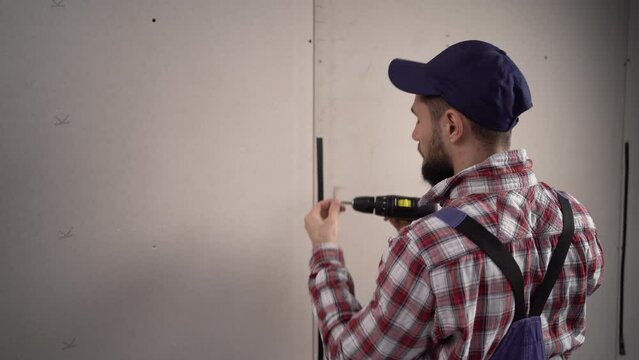Worker builder in blue overalls installs plasterboard drywall at a construction uses electric screwdriver.