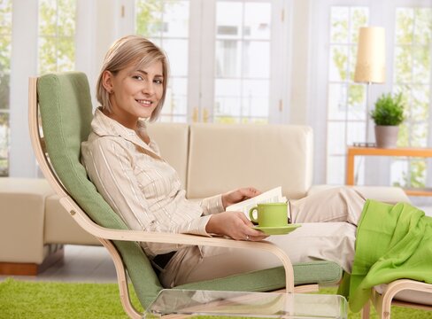 Woman relaxing in armchair at home, reading book , holding coffee cup.