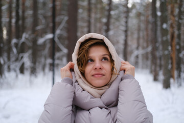 A young woman stands against a background of fir trees under a snowy forest in winter. A girl in a gray down jacket with a hood stands and looks at the camera. Close-up. © ekaterinabyuksel