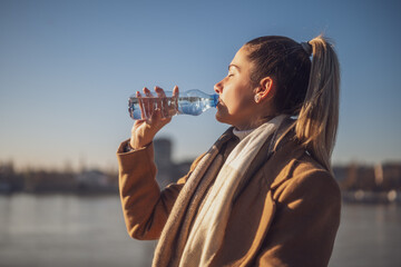 Woman enjoys drinking water while sitting by the river on a sunny winter day.	 Toned image.