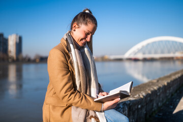 Beautiful woman in warm clothing enjoys  reading book and resting by the river on a sunny winter day. 